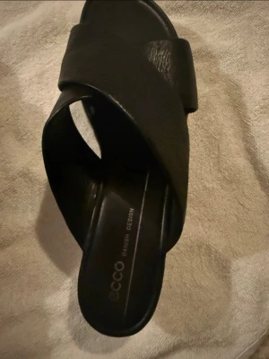 Svarta lädersandaler från Ecco - Stilrena svarta sandaler från Ecco med breda korslagda remmar i mjukt läder. Klassisk slip-in modell med öppen tå och diskret logga på innersulan. Perfekta för en avslappnad och trendig look.