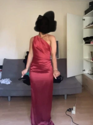 Vinröd one shoulder långklänning från Babyboo - Säljer en vinröd långklänning i satin från Babyboo. Klänningen har en elegant one shoulder-design och öppen rygg. Snygg figurnära passform som framhäver siluetten. Perfekt för speciella tillfällen.