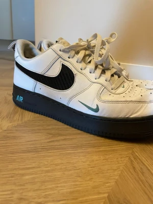 Nike Air Force 1 vit/svart sneaker - Nike Air Force 1 sneakers med vit ovandel i läder, svart mesh Swoosh och svart sula. Grön mini-Swoosh på sidan och AIR-text på hälen. Klassisk låg modell med perforerad tå och vita snören. Perfekt för dig som vill ha en stilren och ikonisk sneaker.