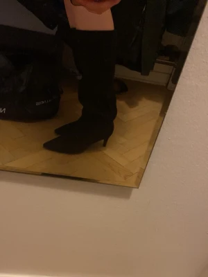 Svarta knähöga boots med klack - Säljer ett par svarta knähöga stövlar med spetsig tå och smal klack. Stövlarna har en stilren design och är tillverkade i ett mockaliknande material. Perfekta för dig som vill ha en edgy och trendig look.