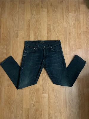 Mörkblå slim fit jeans från Nudie - Säljer ett par mörkblå slim fit jeans från Nudie Jeans med klassisk femficksmodell och snygga slitningar framtill. Jeansen har en diskret logga på bakfickan och är tillverkade i mjuk denim med stretch för extra komfort. Storlek W31 L28, hör av dig vid minsta fundering!