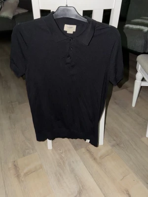 Svart piké från Pull&Bear - Klassisk svart pikétröja från Pull&Bear med korta ärmar och knappar framtill. Tillverkad i mjukt material som känns skönt mot huden. Perfekt till jeans eller shorts för en clean look.