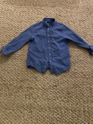 Blå linne skjorta från Ralph Lauren - Klassisk blå linne skjorta från Ralph Lauren med button-down krage och broderad logga på bröstet. Skjortan har långa ärmar och knappar framtill. Perfekt till en avslappnad men ändå stilren look. I storlek 10-12 barn-M