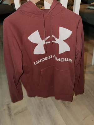 Vinröd hoodie från Under Armour - Säljer en vinröd hoodie från Under Armour med stor vit logga och text på bröstet. Hoodien har huva med dragsko och en klassisk magficka. Tillverkad i mjukt material som känns skönt mot huden. Perfekt för dig som gillar sportig och avslappnad stil.