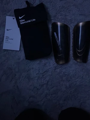Nike Mercurial Lite benskydd svart - Säljer ett par Nike Mercurial Lite benskydd i svart med vit logga och är i L så de kan passar en person i längd 170-180. Lätta och smidiga skydd som ger bra komfort och skydd vid fotbollsträning eller match. Kommer med tillhörande svart förvaringspåse. Perfekt för dig som vill ha stilrena och funktionella benskydd.