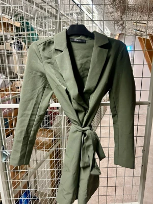 Olivgrön kappa från Gina Tricot - Stilren olivgrön kappa/kavaj från Gina Tricot med knytband i midjan och klassisk krage. Kappan har en rak passform och långa ärmar, perfekt för en trendig och avslappnad look. Materialet känns mjukt och följsamt.