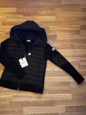 Moncler Cardigan  - Snygg svart dunjacka från Moncler med huva och dragkedja framtill. Jackan har Moncler-logga på ärmen och är tillverkad i ett glansigt material med quiltad design. Perfekt för kalla vinterdagar och har en modern, sportig look.