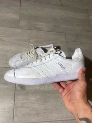 Vita Adidas Gazelle sneakers - Klassiska vita Adidas Gazelle sneakers med lågt skaft och ikoniska tre ränder på sidan. Skorna är tillverkade i mocka och har texten 'GAZELLE' i silver på sidan. Perfekta för dig som gillar stilrena och tidlösa sneakers.