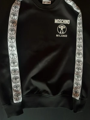 Svart Moschino Milano hoodie - Snygg svart hoodie från Moschino med vita logotexter och emblem på bröstet. Har vita band med Moschino-symboler längs ärmarna för en extra streetstyle-look. Tillverkad i ett glansigt material som känns lyxigt och trendigt. Perfekt för dig som vill sticka ut.