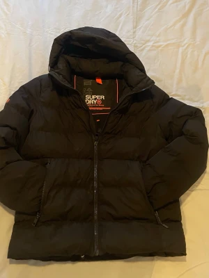 Svart pufferjacka från Superdry - Säljer en svart pufferjacka från Superdry Sports Puffer Series. Jackan har huva, dragkedja framtill och två sidofickor med dragkedja. Tjock och vadderad design som håller dig varm under kalla dagar. Snygga orange detaljer på insidan och klassisk Superdry-logga.