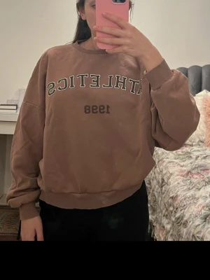 Brun sweatshirt med tryck 'ATHLETICS 1998' - Säljer en oversized brun sweatshirt med texten 'ATHLETICS 1998' i vitt och svart tryck på bröstet. Tröjan har rund hals, ribbade muddar vid ärmslut och nederkant samt en avslappnad passform. Perfekt för en chill och sportig look.