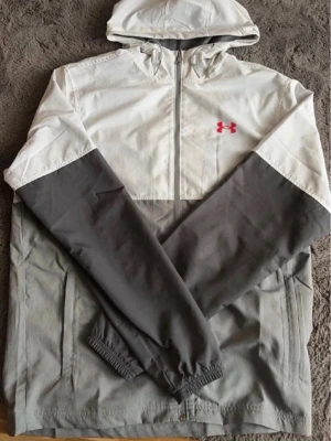Gråvit vindjacka från Under Armour - Snygg vindjacka från Under Armour i grått och vitt med dragkedja framtill och huva. Jackan har elastiska muddar vid ärmsluten och två sidofickor. Under Armour-logga i rött på bröstet. Perfekt för blåsiga dagar och sportiga outfits.