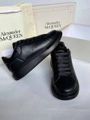 Svarta Alexander McQueen sneakers - Stilrena helsvarta sneakers från Alexander McQueen i slät läder med svarta skosnören och diskret logga på plösen. Klassisk chunky sula och minimalistisk design som passar till det mesta. Perfekta för dig som gillar exklusiva och trendiga skor.
