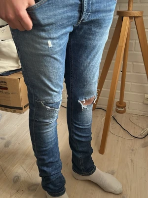 Dondup jeans - Säljer ett par blå skinny jeans med slitningar och hål på knäna. Jeansen har klassisk femficksdesign och dragkedjedetaljer på bakfickorna. Perfekta för en avslappnad och trendig look.