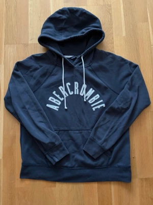 Mörkblå hoodie från Abercrombie - Mörkblå hoodie från Abercrombie med stor vit logga broderad över bröstet. Storlek L sitter mer som en S eller en liten M Skick 7/10, Pris är såklart diskuterbart!