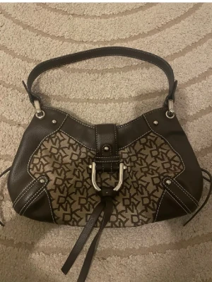 Brun DKNY axelremsväska med monogram - Snygg axelremsväska från DKNY i brunt konstläder och textil med klassiskt DKNY-monogram. Väskan har silverfärgade metalldetaljer, dekorativt spänne framtill och vita kontrastsömmar. Perfekt storlek för det viktigaste och en cool detalj till din outfit. Superfin❤️