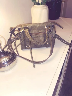 Olivgrön handväska med flätade detaljer - Kompakt olivgrön väska med flätade handtag och guldig dragkedja. Har även justerbar axelrem och ytterficka med dragkedja. I fint skick, och mellan liten