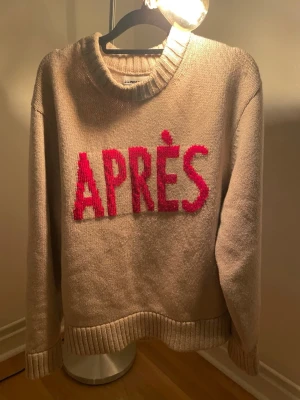 J.Lindeberg (Aprés)  - Stickad beige tröja från J.Lindeberg  med stor röd text 'APRÈS' framtill. Tröjan har rund halsringning, ribbade muddar och avslut. Perfekt för dig som gillar statement-plagg och vill ha något mysigt och trendigt.
