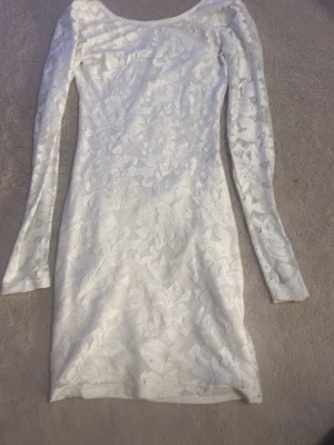 Vit spetsklänning med lång ärm - Snygg vit kortklänning med långärmad design och vackert blommigt spetsmönster. Klänningen har en rundad ringning och figurnära passform, perfekt för dig som vill ha en elegant och stilren look. Materialet är mjukt och följsamt.