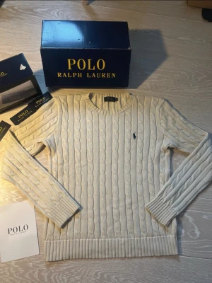 Kabelstickad tröja Polo Ralph Lauren - Kabelstickad tröja i offwhite från Polo Ralph Lauren med rund halsringning och den klassiska broderade loggan på bröstet. Tröjan har långa ärmar och ribbade muddar vid ärmslut och nederkant. Perfekt för en stilren och tidlös look.