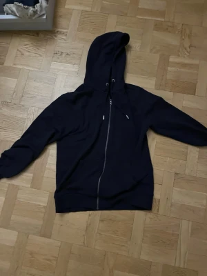 Mörkblå zip hoodie med huva - En stilren mörkblå hoodie med dragkedja framtill och justerbar huva med snören. Hoodien har långa ärmar, ribbade muddar och är tillverkad i ett mjukt material som passar perfekt för en avslappnad look. Inga synliga loggor eller tryck.