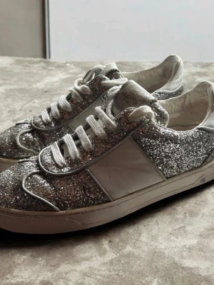 Glittriga Valentino flycrews   - Säljer ett par riktigt snygga sneakers från Valentino flycrew med silverglittrig ovandel, vita detaljer och vita skosnören. Skorna har en låg siluett och cool sula med kamouflagemönster undertill. Perfekta för dig som vill sticka ut med stil. Limmade på vissa ställen samt 4 spikas saknas, skriv vid eventuella frågor 