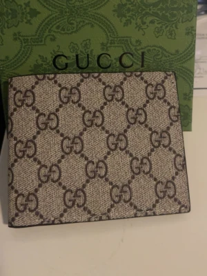 G GG canvas plånbok - Snygg beige och brun plånbok från GG med klassiskt GG-monogram över hela utsidan. Insidan är svart med flera kortfack, ett fönster för ID och präglad GGi-logga. Tillverkad i canvas och läderdetaljer. Perfekt för dig som gillar lyxiga accessoarer.