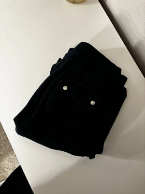 Mörkblå velourbyxor Juicy Couture - Säljer ett par mörkblå mjukisbyxor från Juicy Couture i mjuk velour. Byxorna har bred resår i midjan, fickor fram och en ficka bak med silverfärgade knappar. Perfekta för en chill och avslappnad stil.