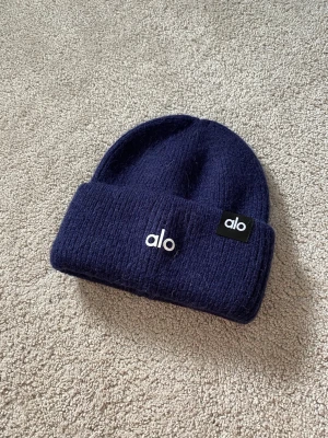 Alo Yoga Mohair/Alpacka Beanie - Snygg mörkblå ribbstickad mössa från Alo med uppvikt kant och vit broderad logga framtill. Mössan har även en svart patch med Alo-loggan på sidan. Perfekt accessoar för kalla dagar och enkel att matcha med olika outfits.