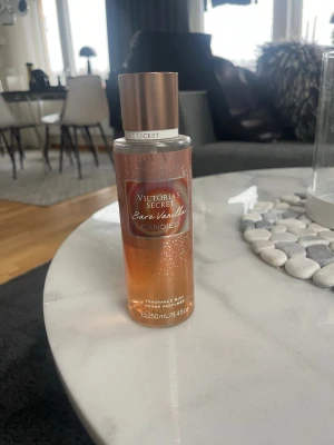 Victoria's Secret Bare Vanilla Candied - En snygg parfymmist från Victoria's Secret i doften Bare Vanilla Candied. Flaskan är cylinderformad i plast med en skimrande kopparfärgad kork och transparent kropp som visar den ljusbruna vätskan. Rymmer 250 ml och har en elegant etikett framtill.