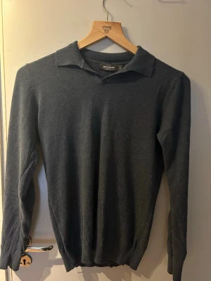 Mörkgrå stickad tröja med krage - Mörkgrå stickad tröja från Dressmann med klassisk krage och långa ärmar. Tröjan har ribbade muddar vid ärmslut och nederkant, vilket ger en snygg och enkel look. Perfekt för dig som gillar stilrena plagg i mjukt material.