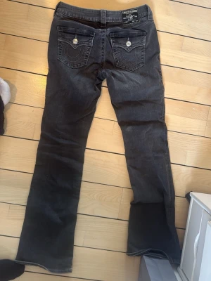 Svarta bootcut jeans True Religion - Säljer ett par svarta bootcut jeans från True Religion med klassiska kontrastsömmar och snygga fickdetaljer bak med silverknappar. Jeansen har låg midja och är tillverkade i denim med lätt stretch. Perfekta för en avslappnad men trendig look.