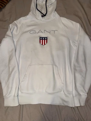 Vit hoodie från GANT - Vit hoodie från GANT med broderad logga och amerikanskinspirerat sköldmärke på bröstet. Klassisk känguruficka framtill och justerbar huva med snören. Tillverkad i mjukt bomullsmaterial som är skönt att ha på sig.