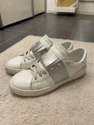Vita och silver sneakers från Valentino - Snygga vita sneakers från Valentino med breda silverdetaljer på sidan och tungan. Skorna har klassisk låg profil, vita snören och guldfärgad logga på plösen. Tillverkade i slätt läder för en clean och exklusiv look. Säljer dom pågrund av att dom är för små för mig!!💓💓