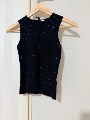 Svart ribbad ärmlös topp med pärlor - Snygg svart ribbad topp utan ärmar, dekorerad med små vita pärlor över hela plagget. Toppen har en hög halsringning och är figurnära i modellen. Perfekt att styla med jeans eller kjol för en trendig look.