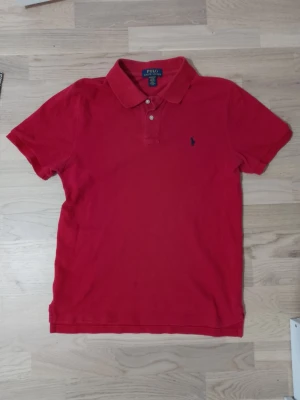 Röd pikétröja från Polo Ralph Lauren - Klassisk röd pikétröja från Polo Ralph Lauren med korta ärmar och krage. Tröjan har två knappar vid halsen och den ikoniska lilla logotypen broderad på bröstet. Tillverkad i mjuk bomull för en bekväm känsla.