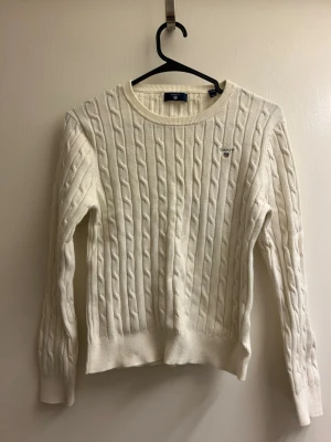 Vit kabelstickad tröja från Gant - Kabelstickad vit tröja från Gant med rund halsringning och diskret logga broderad på bröstet. Tröjan har långa ärmar och ribbade muddar vid ärmslut och nederkant. Klassisk och tidlös design i mjukt material.
