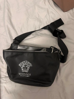 Svart magväska från  Versace Parfums - Snygg svart magväska från Versace Parfums med vit Medusa-logga framtill. Väskan har justerbart band och dragkedja upptill. Tillverkad i ett slätt, tåligt material som ger en clean och modern look. Perfekt för att bära det viktigaste med stil.