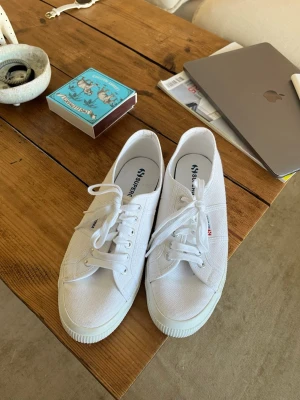 Vita låga sneakers från Superga - Klassiska vita sneakers från Superga i canvas med vita snören och gummisula. Modellen är låg och har diskret Superga-logga på sidan. Perfekta till jeans eller shorts för en clean och stilren look.