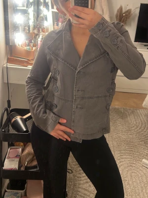 Grå dubbelknäppt rock med detaljer - Snygg grå höst jacka  med dubbelknäppning och stora dekorativa knappar. Jackan har markerad krage, figurnära passform och detaljerade sömmar. Köpt på Sellpy men märket är Pimkie 