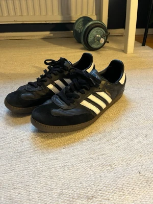 Adidas Samba svarta sneakers - Klassiska Adidas Samba sneakers i svart läder och mocka med vita detaljer. Skorna har bruna gummisulor och svart snörning. 