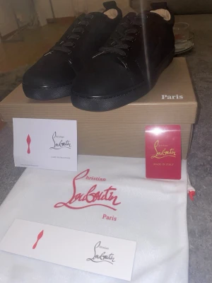 Svarta Christian Louboutin sneakers - Snygga svarta sneakers från Christian Louboutin i mocka med klassisk röd sula och diskreta detaljer. Lågt skaft och svarta skosnören ger en clean look. Perfekta för dig som vill ha något exklusivt och stilrent i garderoben.