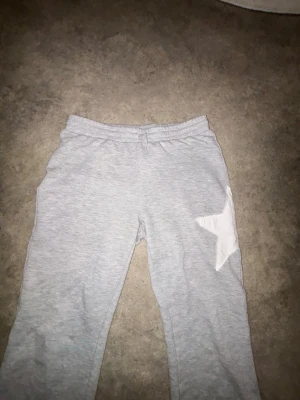 Grå mjukisbyxor med vit stjärna - Säljer ett par ljusgrå mjukisbyxor med resår i midjan och en stor vit stjärna på ena benet. Byxorna har raka ben och är gjorda i mjukt, bekvämt material som passar perfekt för chill dagar hemma.