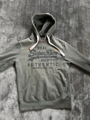 Hoodie från Superdry - Fet  hoodie från Superdry med stor logga och text. Passar perfekt in för våren där man kan rocka en avslappnad stil. Hör av dig om du har några frågor.