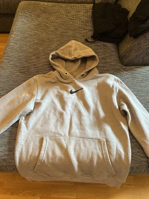 Grå hoodie från Nike med logga - Säljer en grå hoodie från Nike med klassisk svart swoosh-logga på bröstet. Hoodien har stor känguruficka framtill och justerbar huva med dragsko. Tillverkad i mjukt bomullsmaterial som är skönt mot huden. Perfekt för en avslappnad och sportig stil.