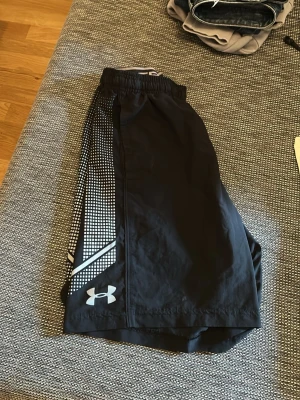 Svarta träningsshorts från Under Armour - Snygga svarta träningsshorts från Under Armour med vitt rutmönster på sidorna och logga framtill. Shortsen har elastisk midja och är tillverkade i ett lätt och snabbtorkande syntetmaterial, perfekta för gymmet eller löprundan.