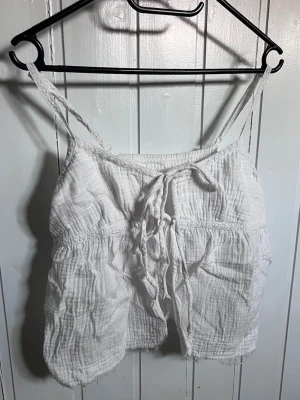 Vit linnetopp med snörning - Säljer en luftig vit linnetopp med tunna virkade axelband och snygg snörning framtill. Toppen har en avslappnad passform och är gjord i ett mjukt, strukturerat tyg som känns perfekt för varma dagar.