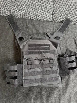 Taktisk väst och benhölster svart - Säljer ett set med taktisk väst och benhölster i svart nylon. Västen har justerbara axelband, MOLLE-system och kardborre för patchar. Benhölstret har remmar för att fästa runt benet och passar till airsoft eller paintball. Perfekt för dig som gillar taktisk utrustning.