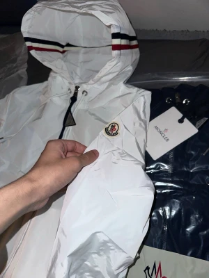 Helt Ny Vit Moncler liknande vindjacka med huva - Snygg vit vindjacka från Moncler med huva och dragkedja framtill. Jackan har Moncler-logga på ärmen och detaljer i rött, vitt och blått på huvan. Tillverkad i lätt och glansigt material, perfekt för blåsiga dagar. Två sidofickor med dragkedja och justerbar huva. Lägg märke till mitt användarnamn