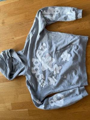 Grå spider hoodie med tryck - Säljer en grå hoodie med stora vita grafiska tryck på bröstet och ärmarna. Hoodien har en klassisk huva och magficka. Tillverkad i mjuk bomull, perfekt för en avslappnad streetwear-look.